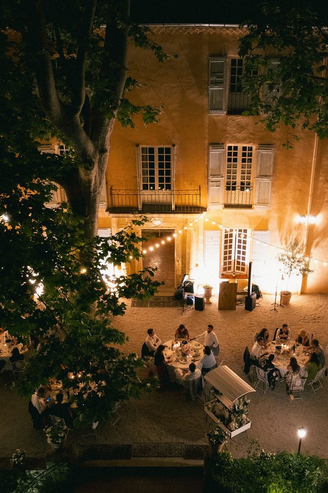 The illuminated façade from above, guests dining on the gravel